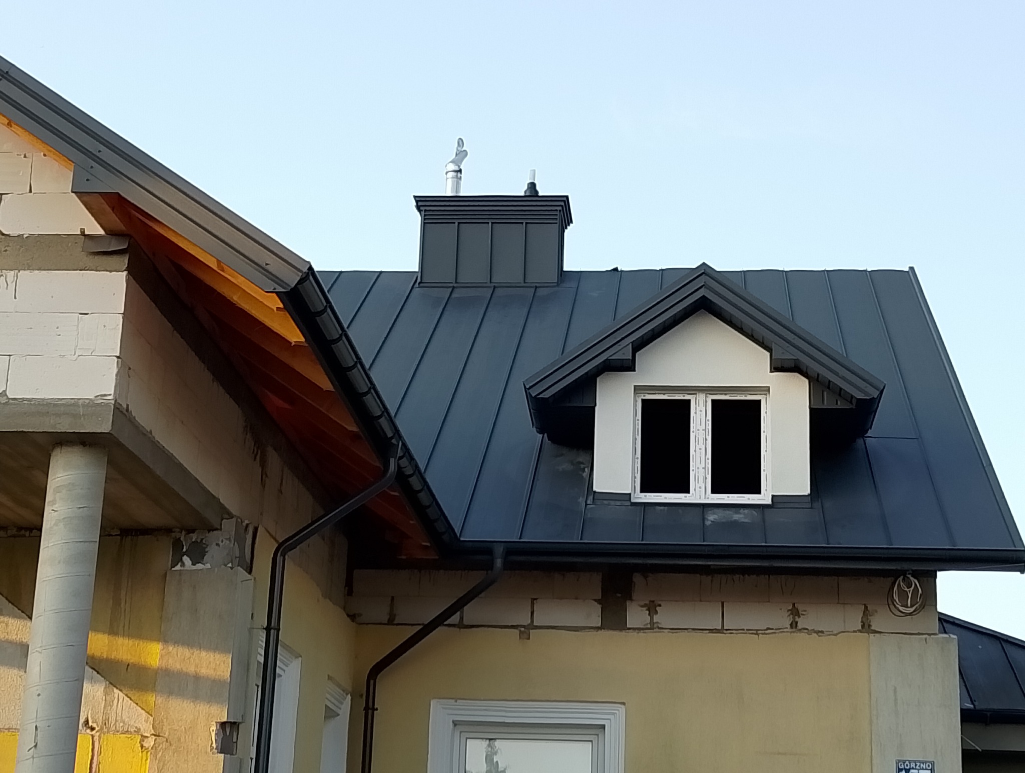 Ciemny dach z blachy na budynku w trakcie budowy, z widocznym kominem i oknem dachowym. Rynny zamontowane, widoczne elementy konstrukcyjne budynku.