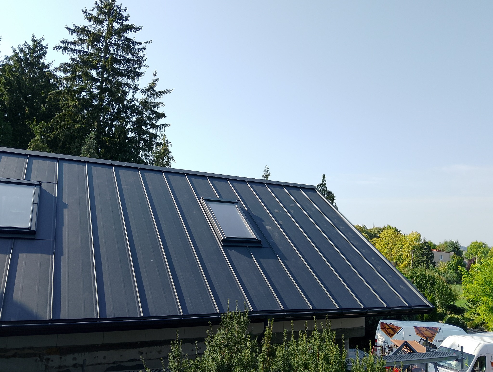 Nowoczesny, ciemny dach z blachy na rąbek stojący z oknem dachowym, na tle zieleni i błękitnego nieba. Widoczny fragment rynny i elewacji budynku.