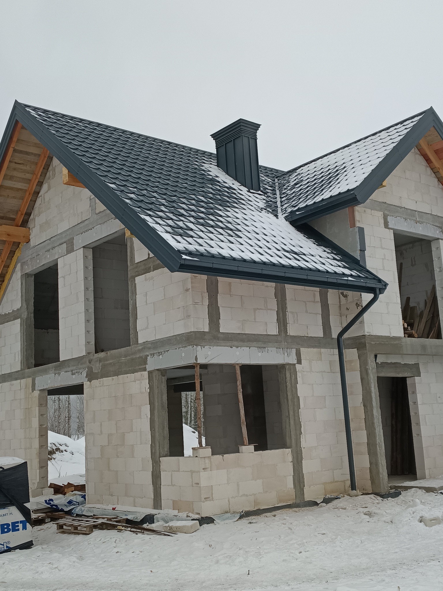 Dom w stanie surowym otwartym, z częściowo wykonanym dachem z blachodachówki w kolorze grafitowym i zamontowaną rynną. Widoczny komin i śnieg.