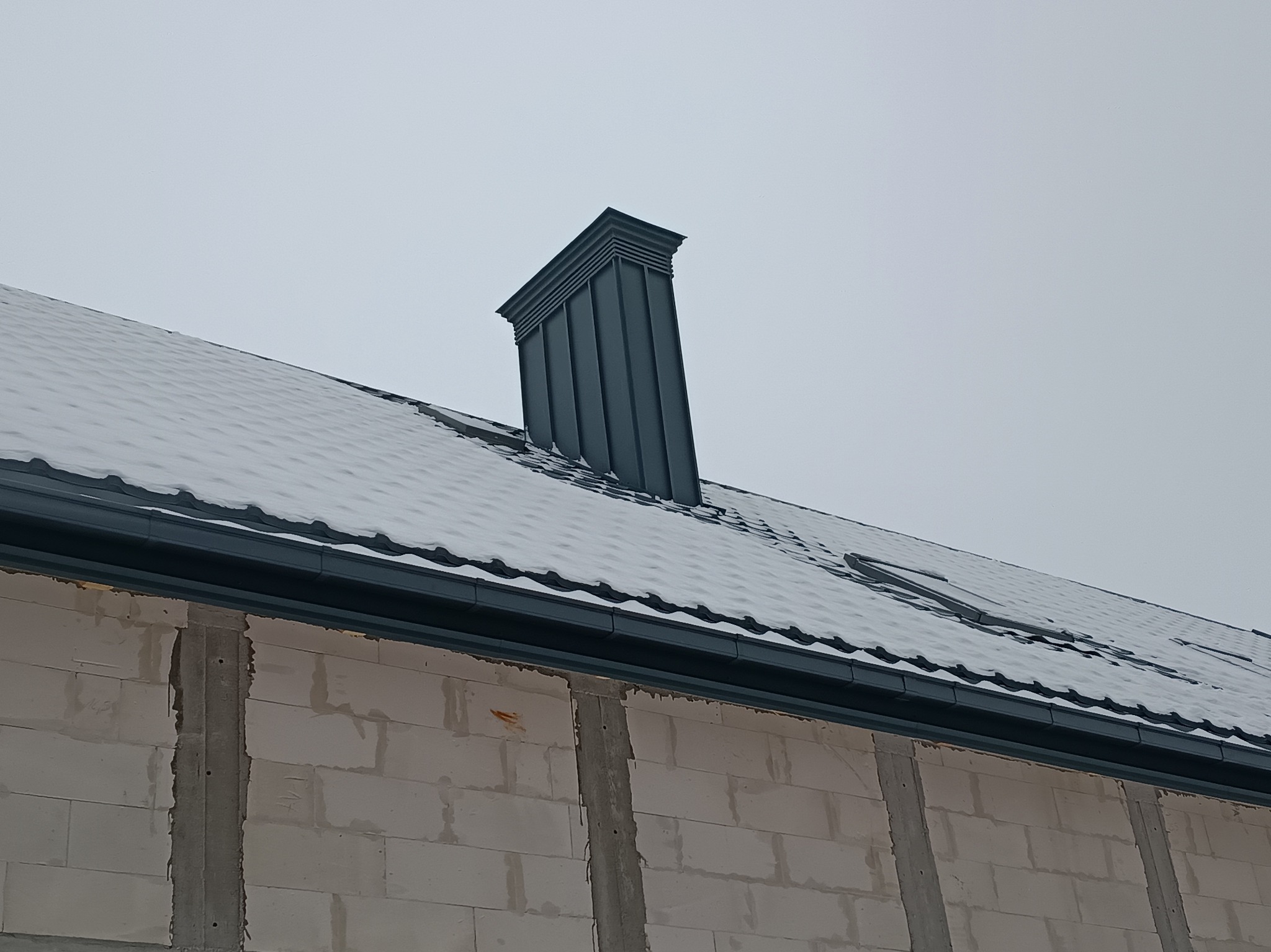 Szary dach pokryty śniegiem z nowoczesnym kominem i rynnami, widoczne ściany budynku z betonu komórkowego. Pochmurne niebo w tle.