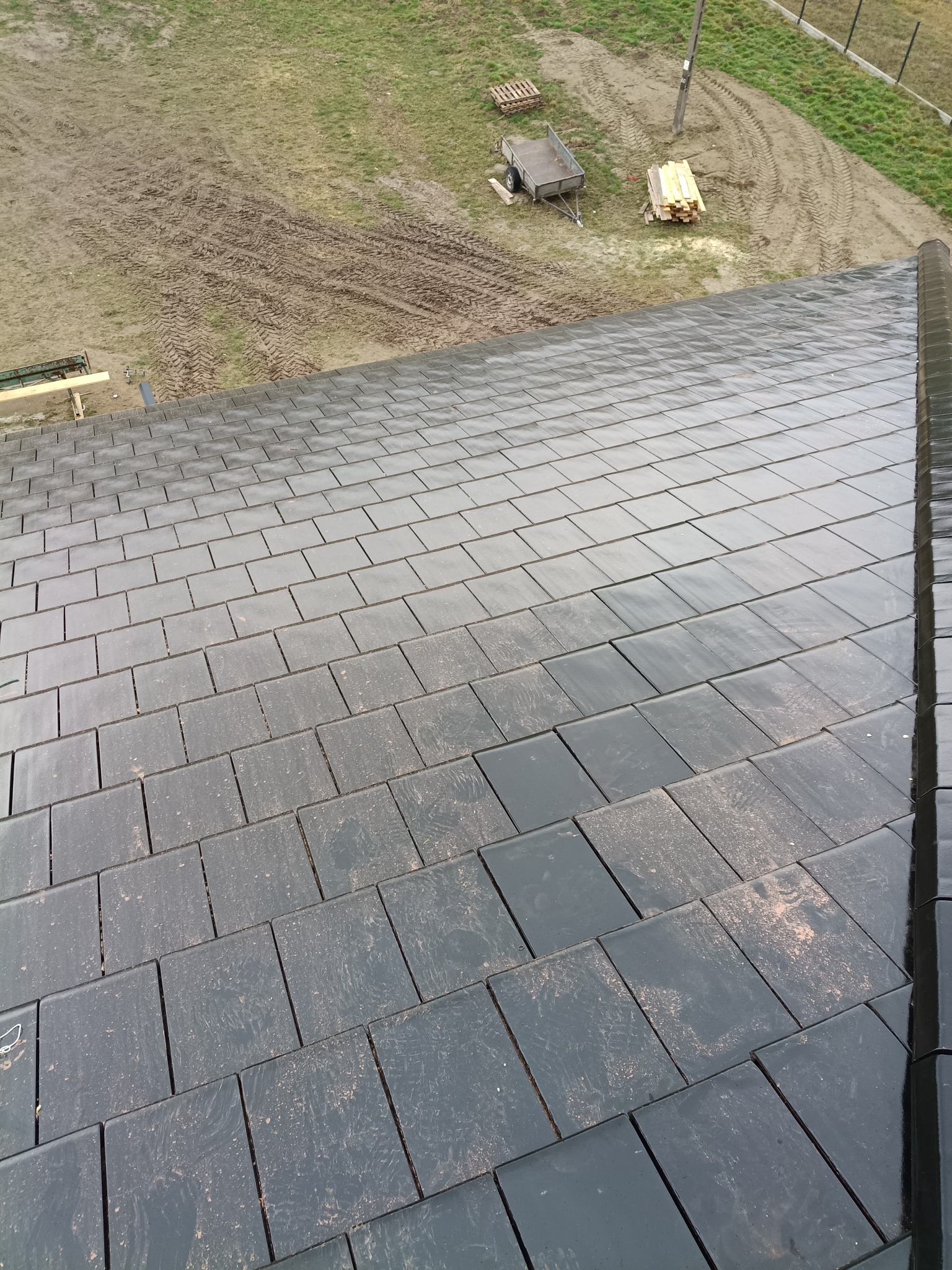 Nowy, błyszczący dach z ciemnej dachówki ceramicznej, widziany z góry pod kątem. W tle widoczne ślady opon na ziemi i przygotowane drewno na paletach.