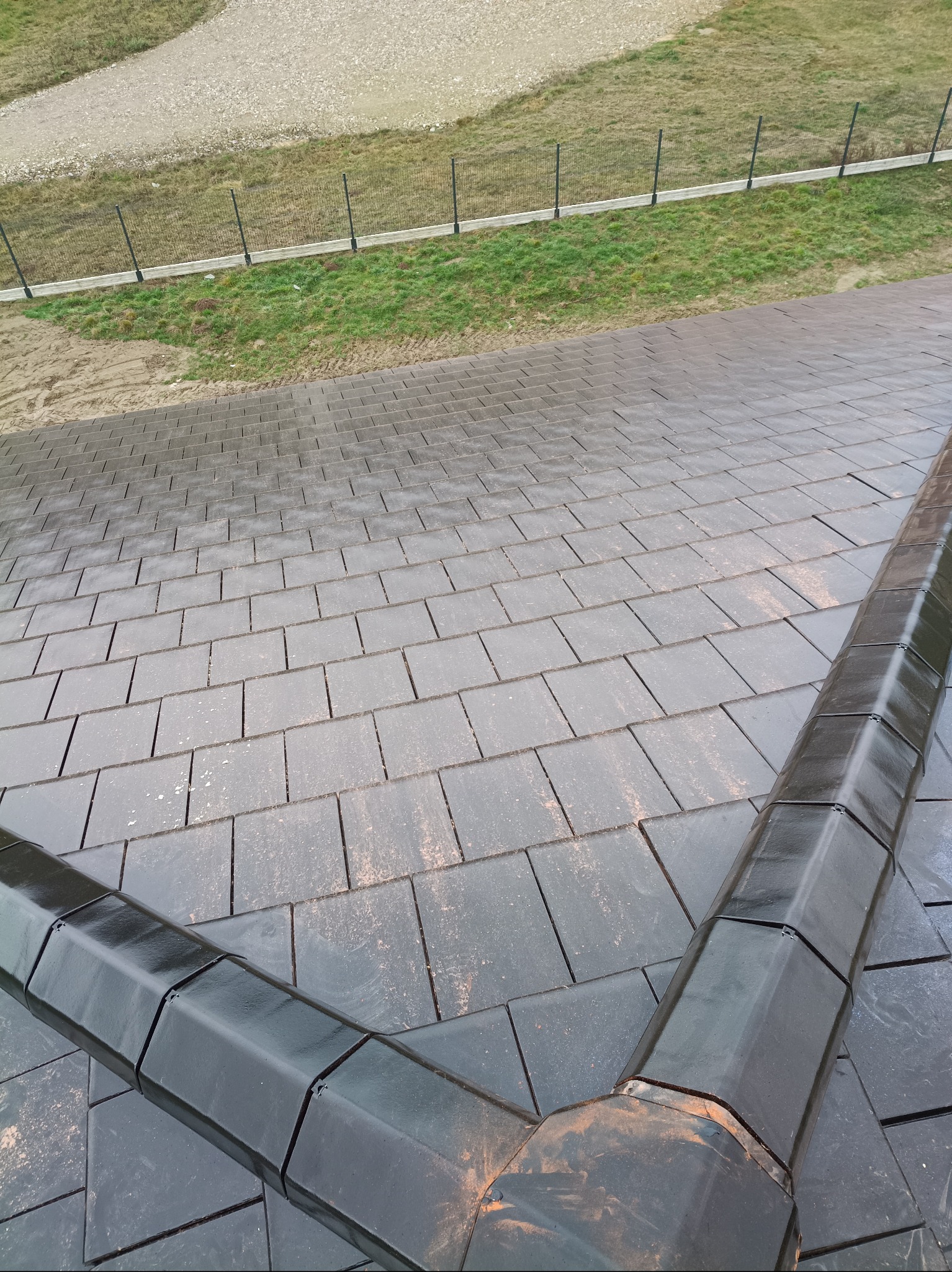 Ukończony dach z ciemnej dachówki ceramicznej, z widocznym systemem odprowadzania wody w narożniku. W tle trawa i ogrodzenie.