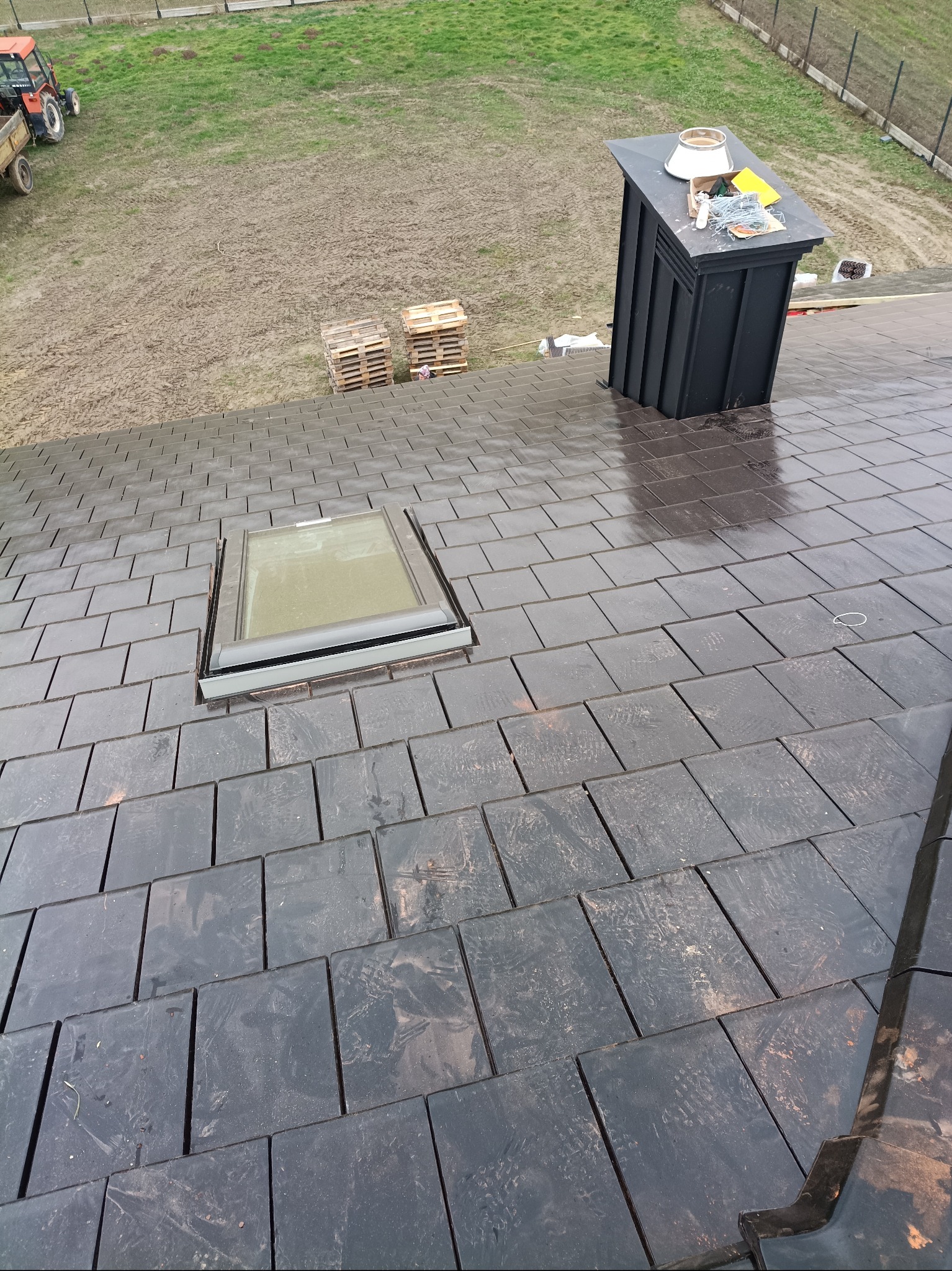 Nowy dach z czarnej dachówki ceramicznej z oknem dachowym i kominem wentylacyjnym. Na dachu widoczne narzędzia i akcesoria dekarskie. W tle traktor i palety.