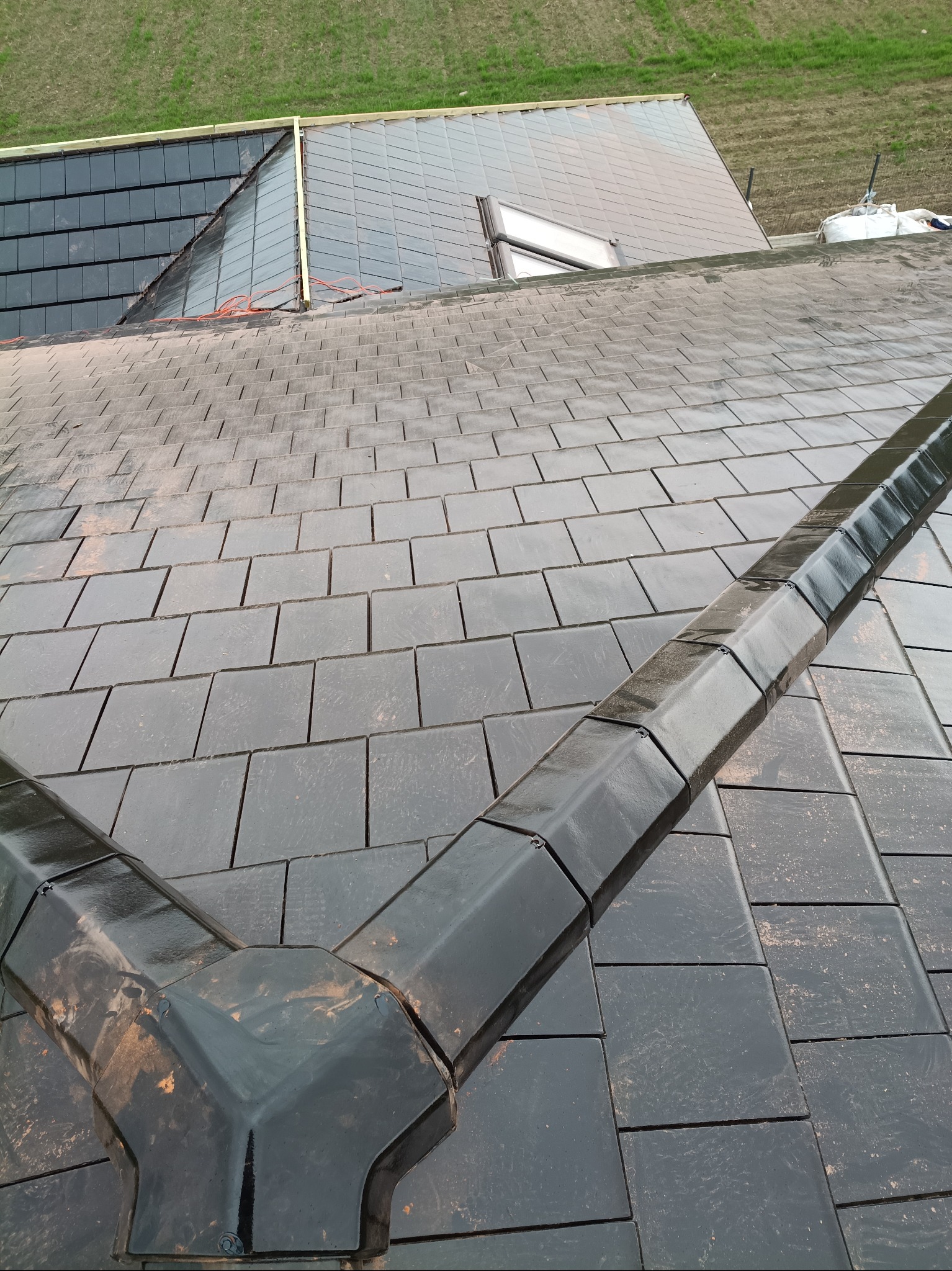 Czarny dach z dachówki ceramicznej, widok z góry na fragment połaci z rynną koszową. W tle fragment dachu z oknem połaciowym i zielone pole.