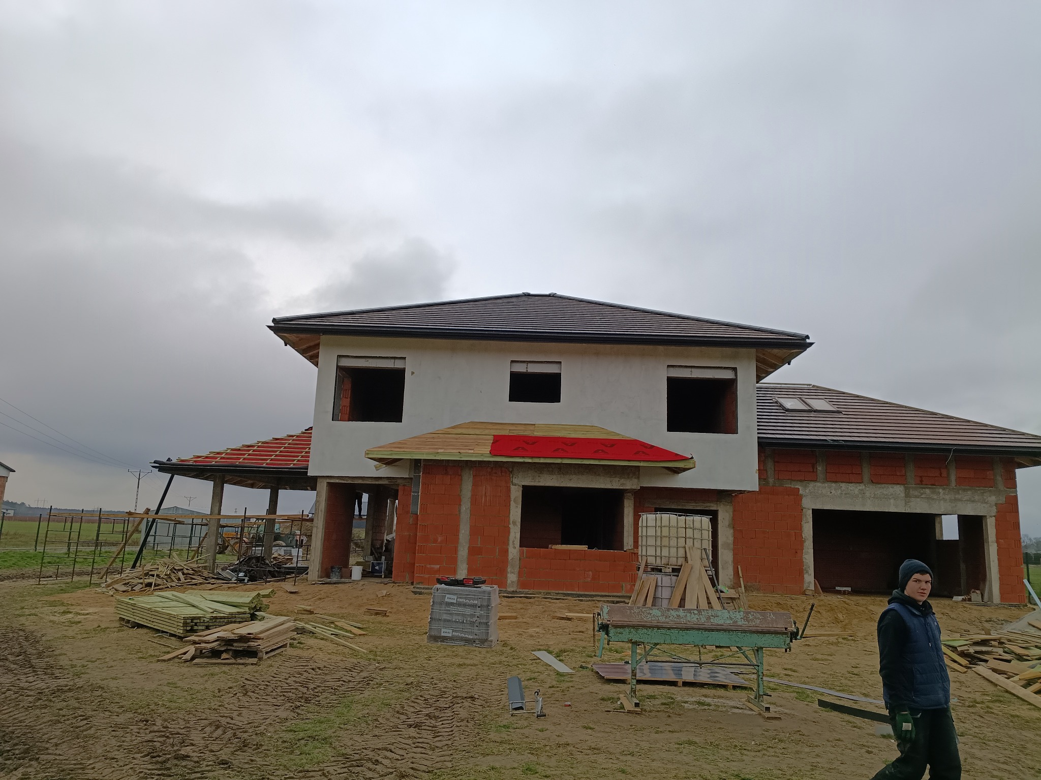 Budowa domu jednorodzinnego w trakcie realizacji. Widoczny fragment dachu z brązową dachówką, ściany z czerwonej cegły i pracownik w ubraniu roboczym.