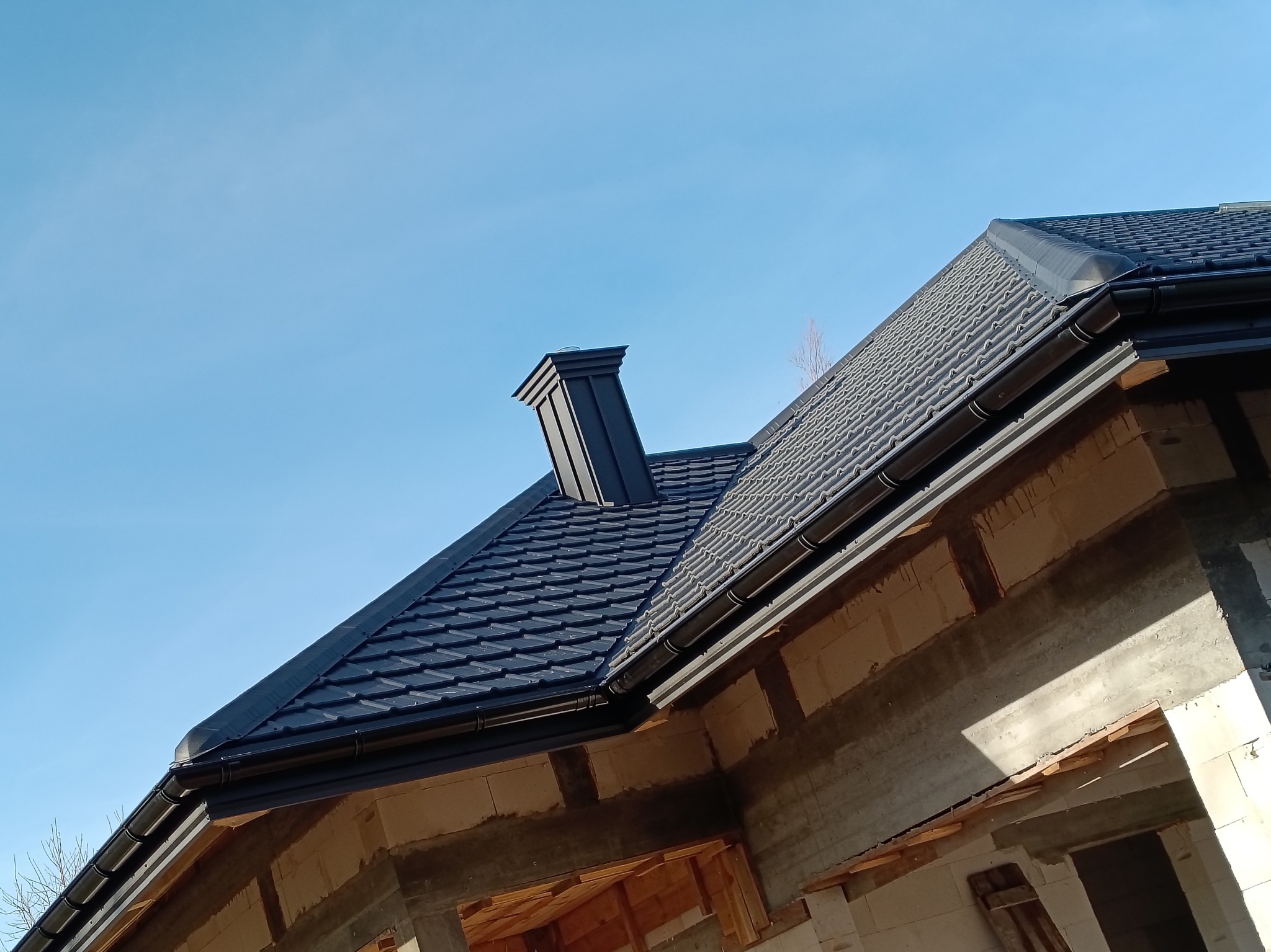 Nowy, grafitowy dach z blachodachówki modułowej i orynnowaniem, widoczny z dołu na tle błękitnego nieba. Budynek w stanie surowym otwartym.