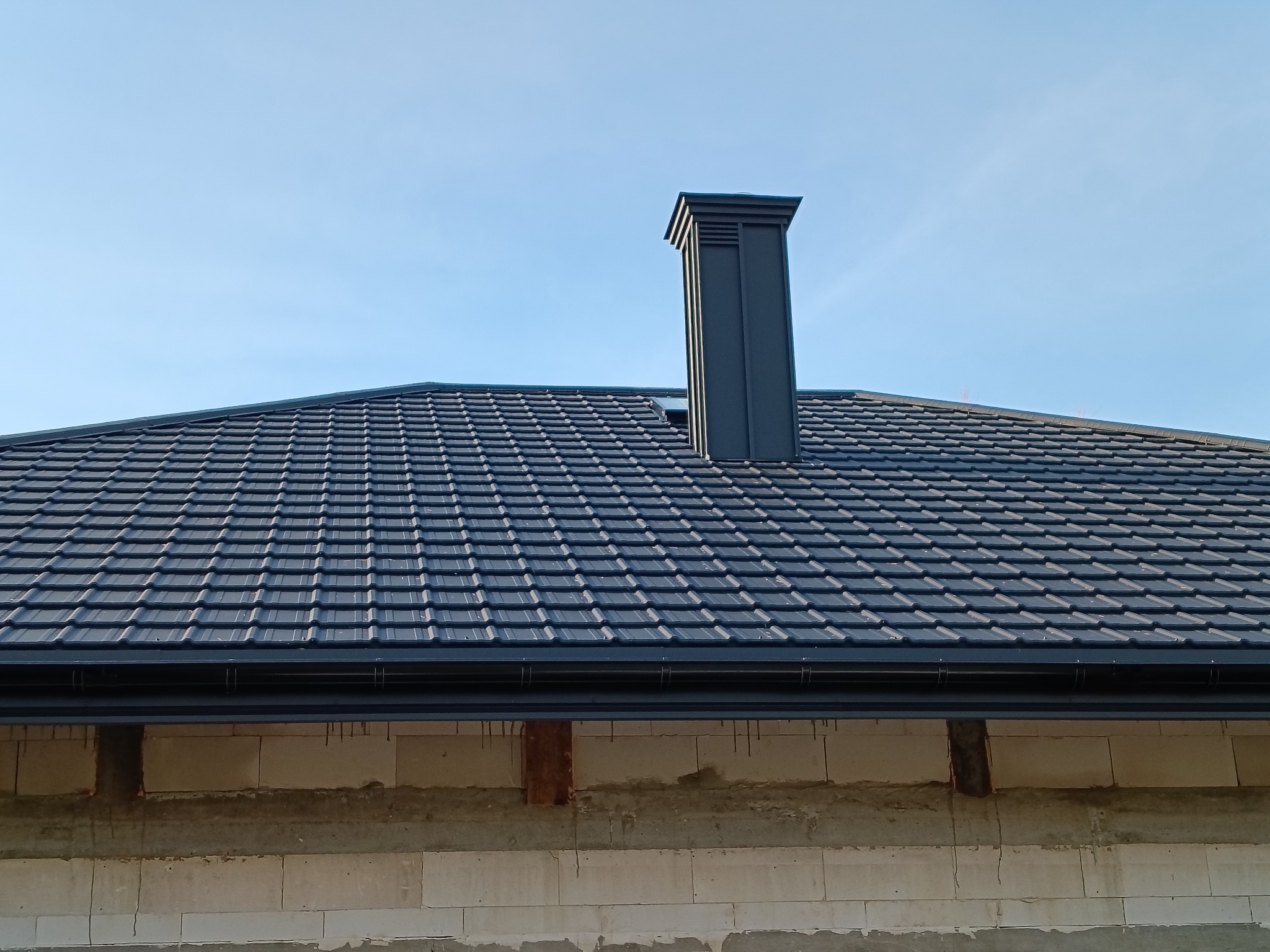 Grafitowy dach z dachówki modułowej i nowoczesny komin na tle błękitnego nieba. Widoczne orynnowanie i fragment ściany budynku z betonu komórkowego.