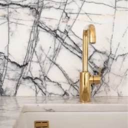 Błyszcząca złota bateria umywalkowa na tle marmurowej ściany z wyraźnym użyleniem, widoczny fragment białej umywalki z minimalistycznym odpływem.