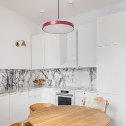 Jasna kuchnia z białymi szafkami, marmurowym backsplashiem i okrągłym drewnianym stołem z krzesłami. Czerwona, okrągła lampa sufitowa. Na blacie misa z owocami.