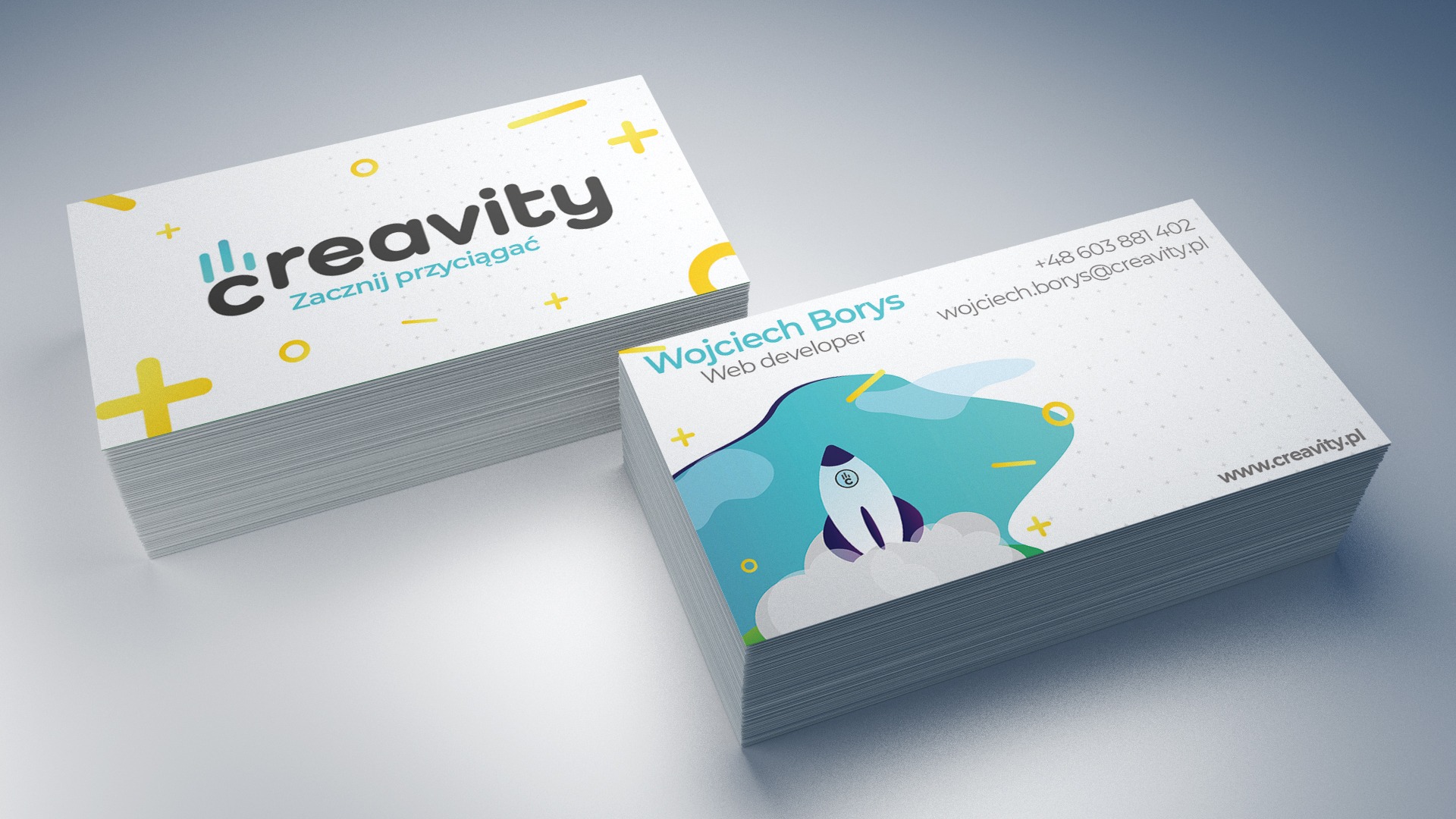 Dwa stosy wizytówek firmy Creavity z motywem rakiety i hasłem 'Zacznij przyciągać', leżące na gładkim, szarym tle. Jedna wizytówka prezentuje dane kontaktowe Wojciecha Borysa, web developera.