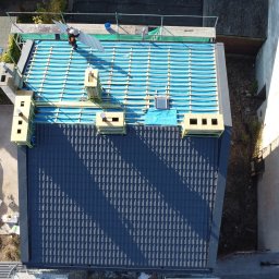 Giera-Bud Mariusz Giera - Widok z góry na dach w trakcie krycia. Część dachu pokryta ciemną dachówką, część z izolacją i łatami. Na dachu dekarz z elementem dachówki.