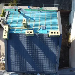 Widok z góry na dach w trakcie krycia. Część dachu pokryta ciemną dachówką, część z izolacją i łatami. Na dachu dekarz z elementem dachówki.