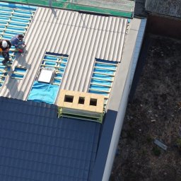 Giera-Bud Mariusz Giera - Układanie dachówki na konstrukcji dachu z widocznymi otworami okiennymi i kominami. Prace dekarskie na dachu z niebieską membraną. Widok z góry.