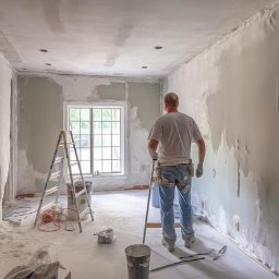 Giera-Bud Mariusz Giera - Remont pokoju: mężczyzna w białej koszulce szpachluje ścianę, drabina, okno, ślady szpachli na podłodze, narzędzia. Widok z tyłu, wnętrze w trakcie prac.