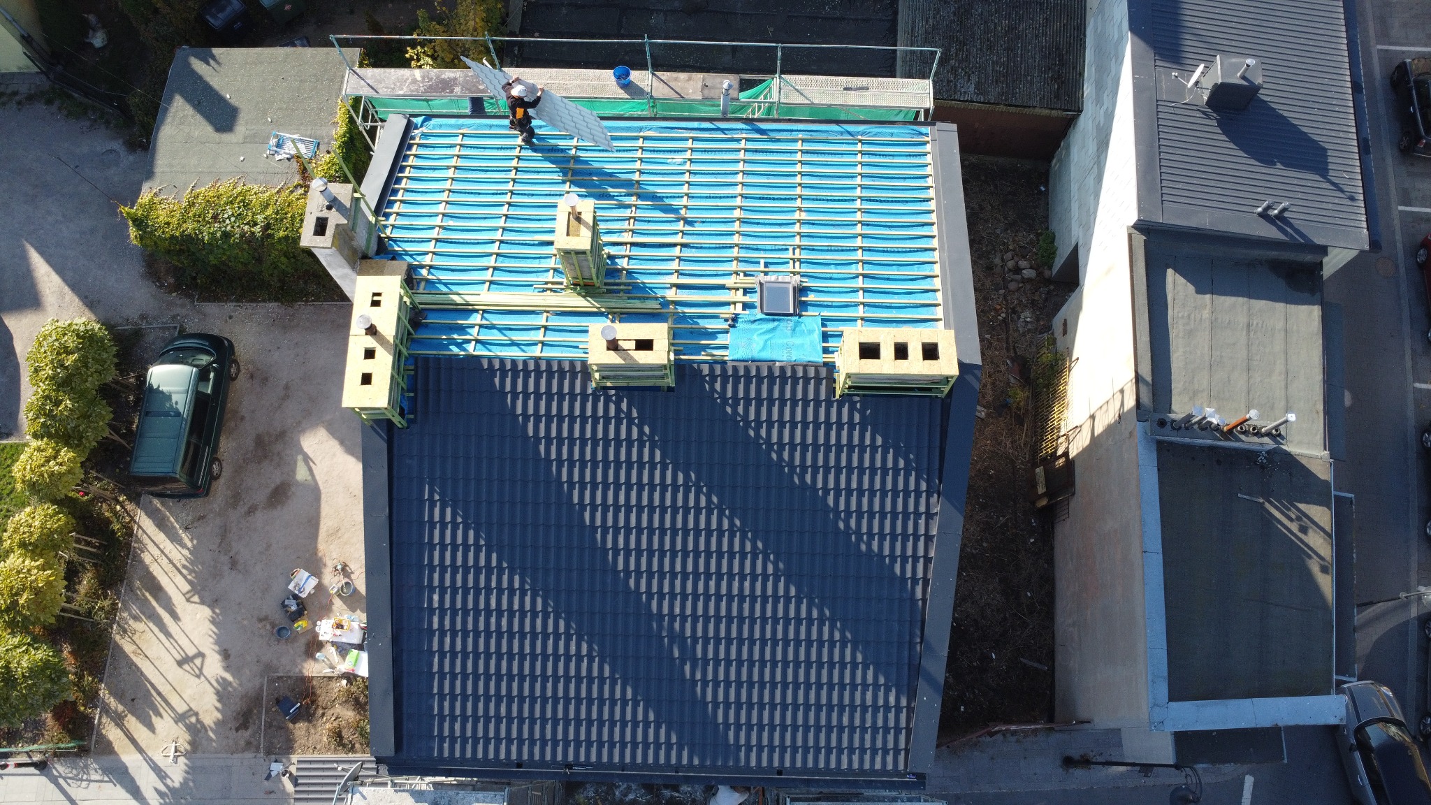 Widok z góry na dach w trakcie krycia. Część dachu pokryta ciemną dachówką, część z izolacją i łatami. Na dachu dekarz z elementem dachówki.