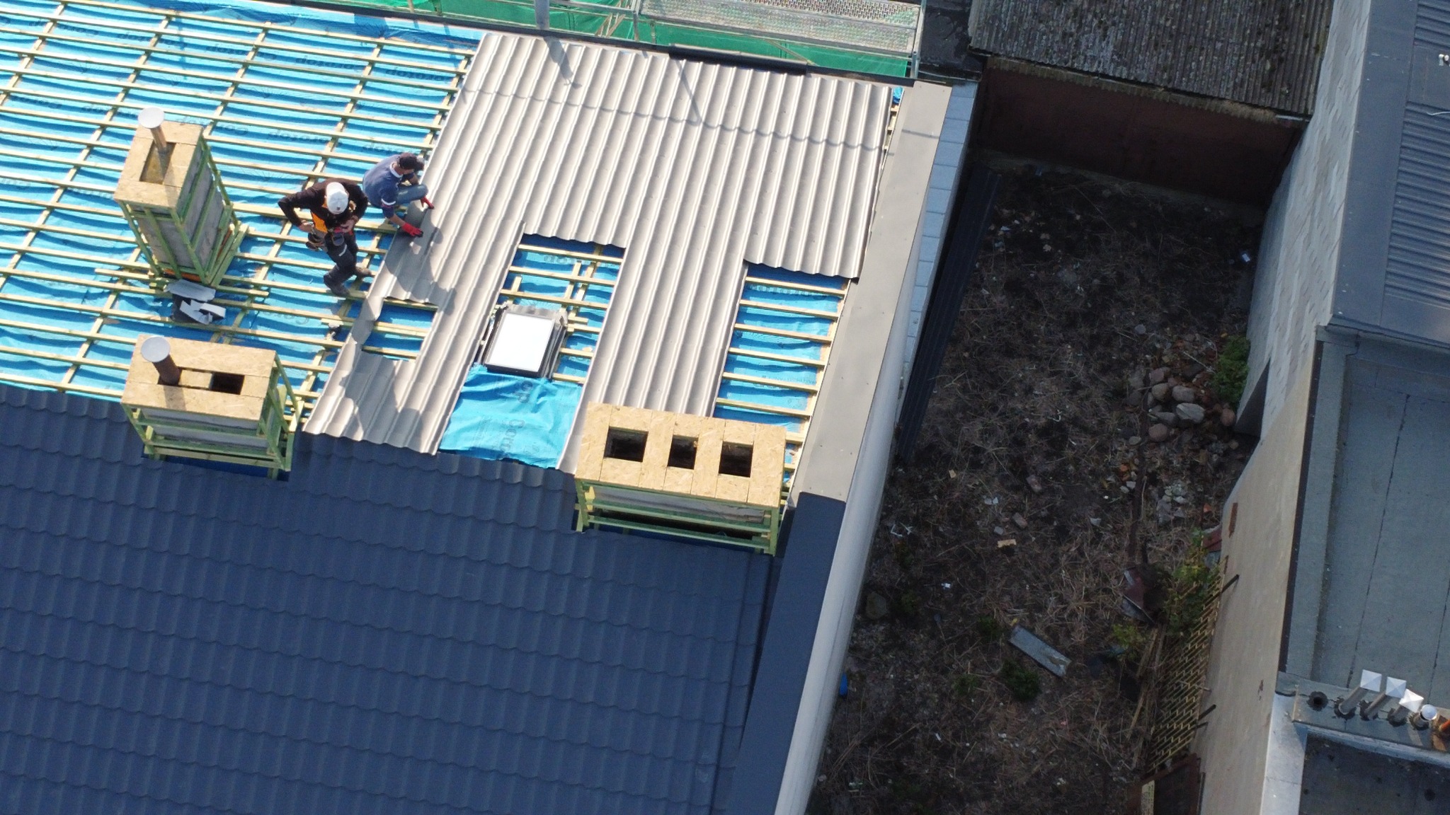 Układanie dachówki na konstrukcji dachu z widocznymi otworami okiennymi i kominami. Prace dekarskie na dachu z niebieską membraną. Widok z góry.