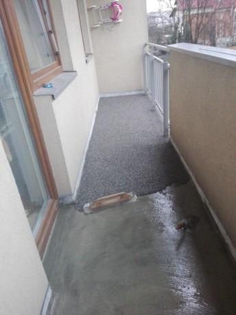 Balkon w trakcie renowacji. Część podłogi pokryta szarym, kamyczkowym wzorem, reszta zalana mokrą wylewką. Widoczna kielnia. Biała balustrada i fragment okna.