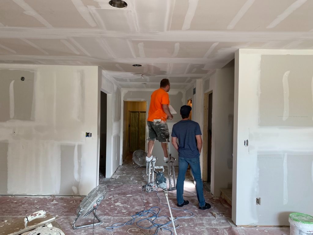 Wnętrze w trakcie prac wykończeniowych; ściany i sufit pokryte płytami gipsowo-kartonowymi, szpachlowane łączenia, dwóch mężczyzn w trakcie pracy, narzędzia na podłodze.