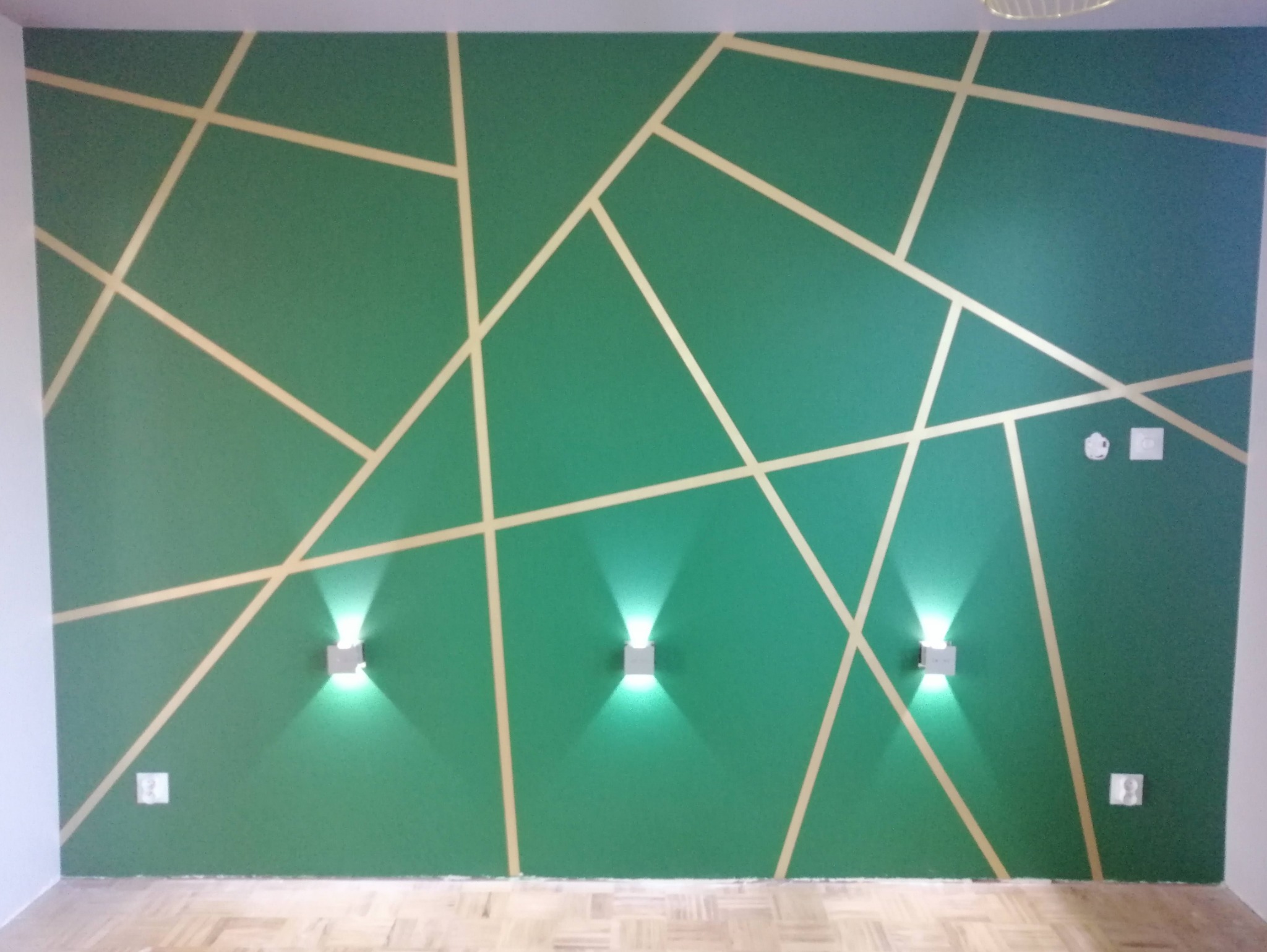Szmaragdowa ściana z geometrycznym wzorem z jasnych pasków, oświetlona trzema nowoczesnymi kinkietami, widoczne gniazdka elektryczne i fragment parkietu.