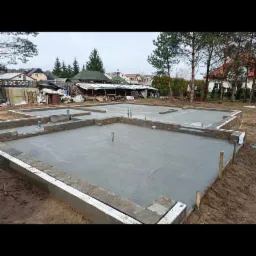 Wylany betonowy fundament pod budynek jednorodzinny, widoczne wystające rury i elementy zbrojeniowe na tle zabudowy wiejskiej.
