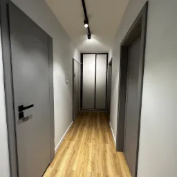 Wnętrze korytarza z panelami podłogowymi imitującymi drewno, szare drzwi po obu stronach i oświetleniem punktowym na suficie.