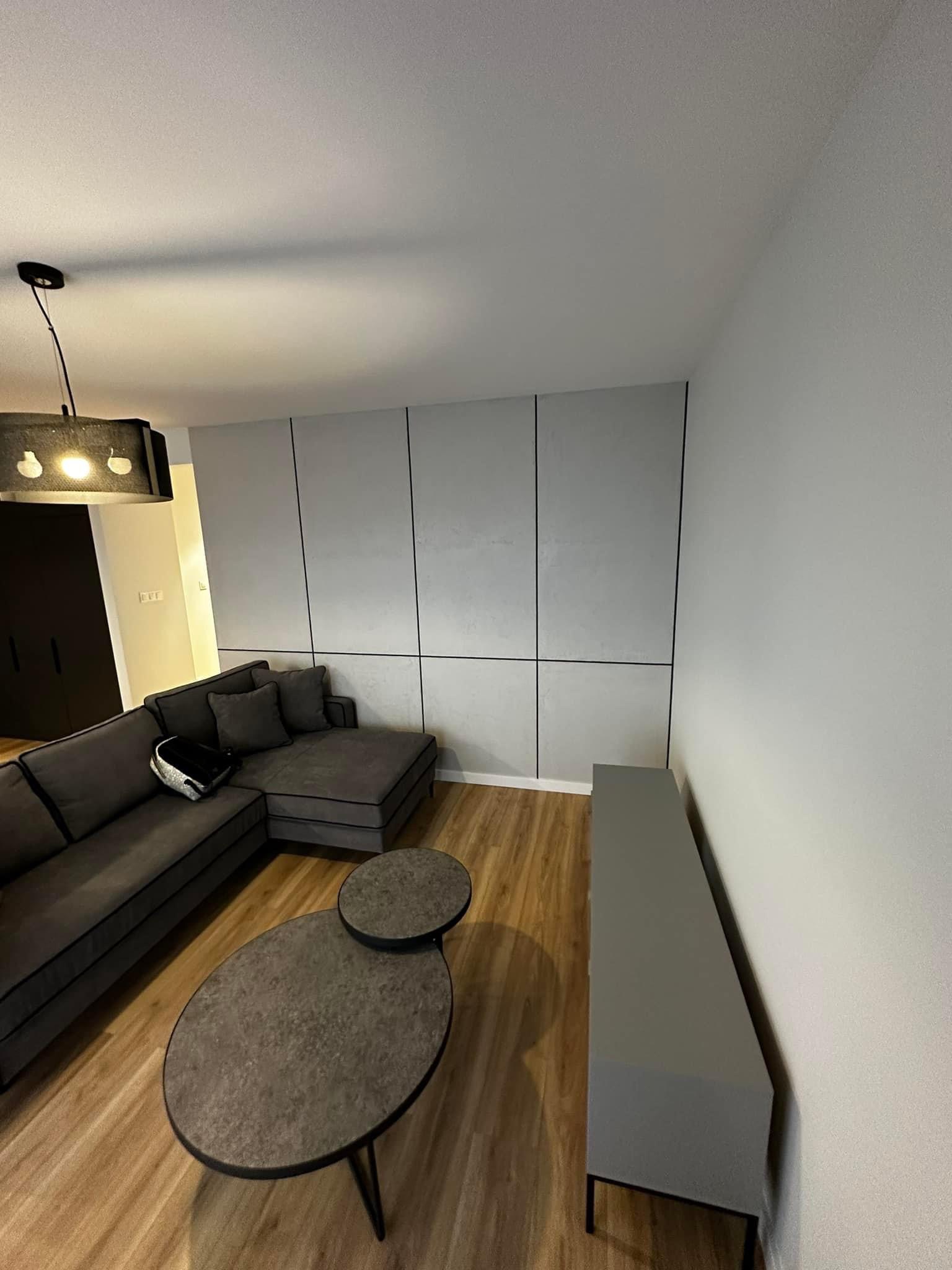 Nowoczesny salon z szarą sofą narożną, stolikami kawowymi o nieregularnym kształcie i ścianą wykończoną panelami w odcieniach szarości z czarnymi listwami.