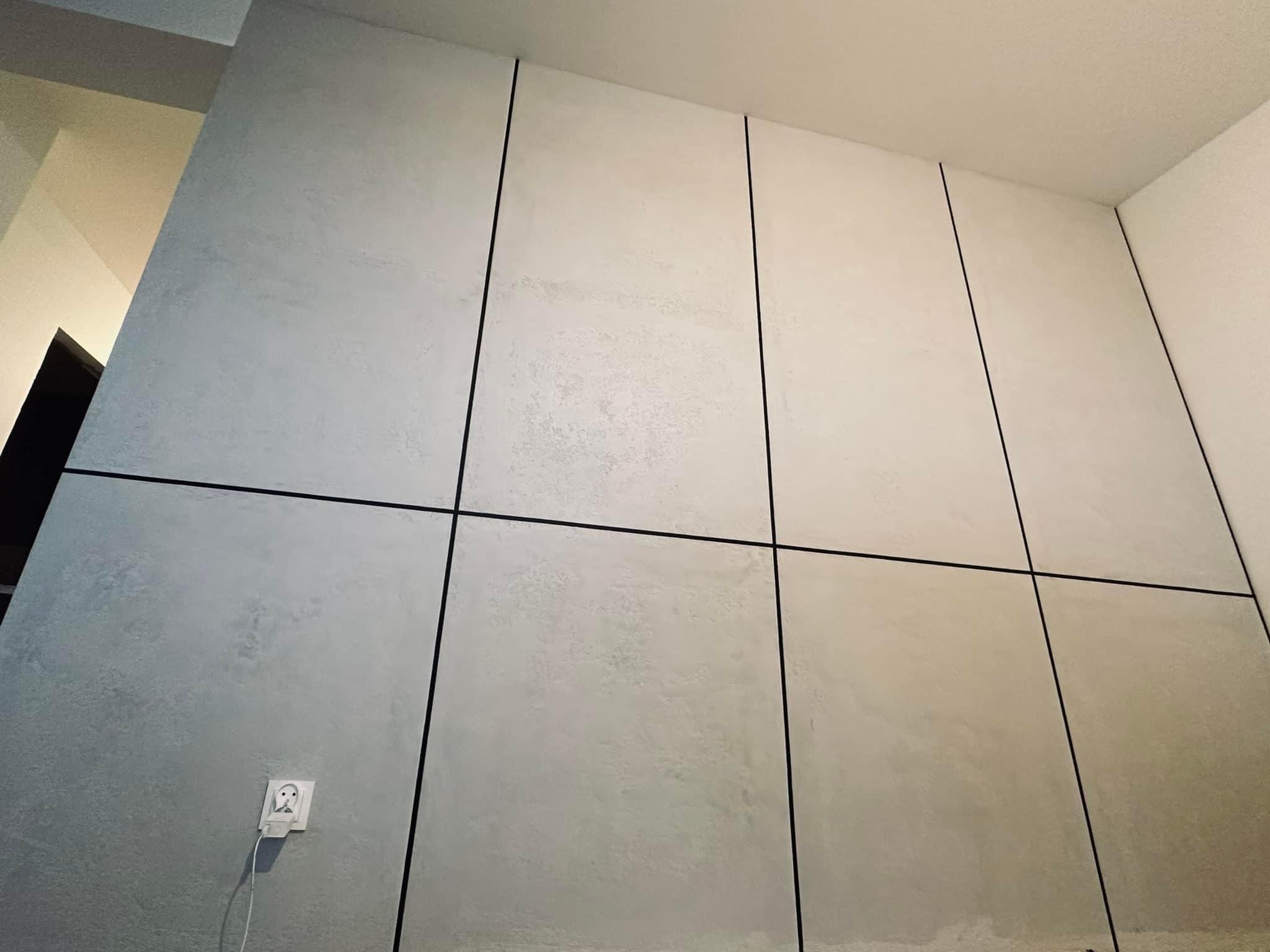 Ściana wykończona dekoracyjną szarą farbą z czarnymi listwami tworzącymi geometryczny wzór; widoczne gniazdko elektryczne.