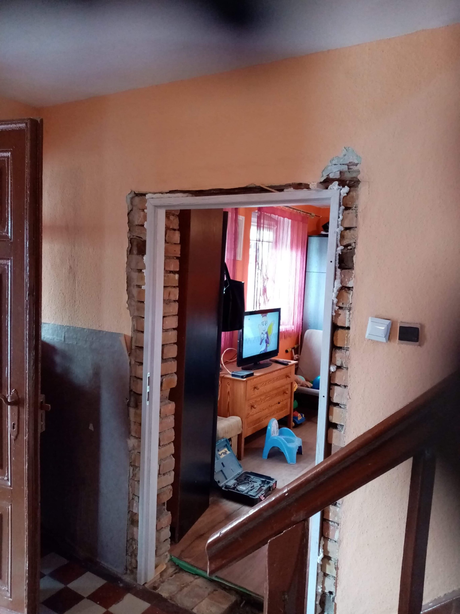 Przygotowanie otworu drzwiowego w ścianie z cegły, widoczne uszkodzenia muru i pianka montażowa, w tle meble i dziecięce zabawki.