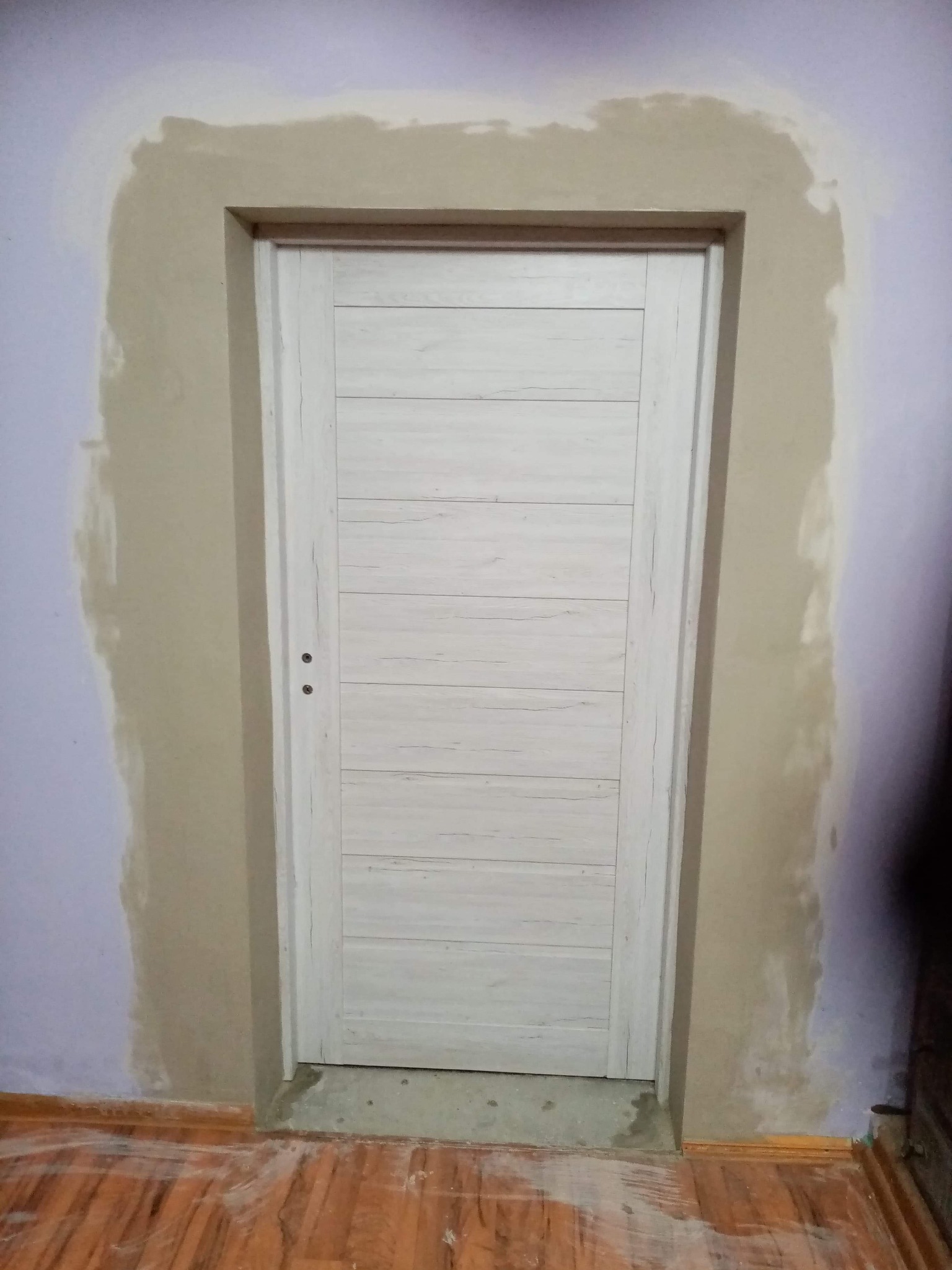 Świeżo zamontowane, białe drzwi wewnętrzne z poziomymi panelami, widoczne ślady gipsu na ścianie i podłodze po montażu ościeżnicy.