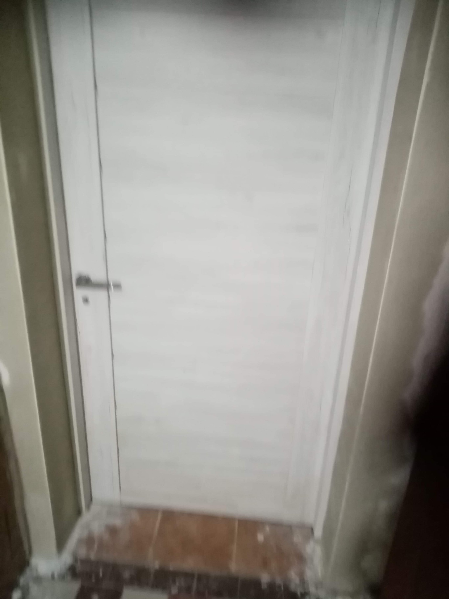 Białe drzwi wewnętrzne z metalową klamką, widoczne ślady uszczelniania wokół ościeżnicy i na brązowych płytkach podłogowych.