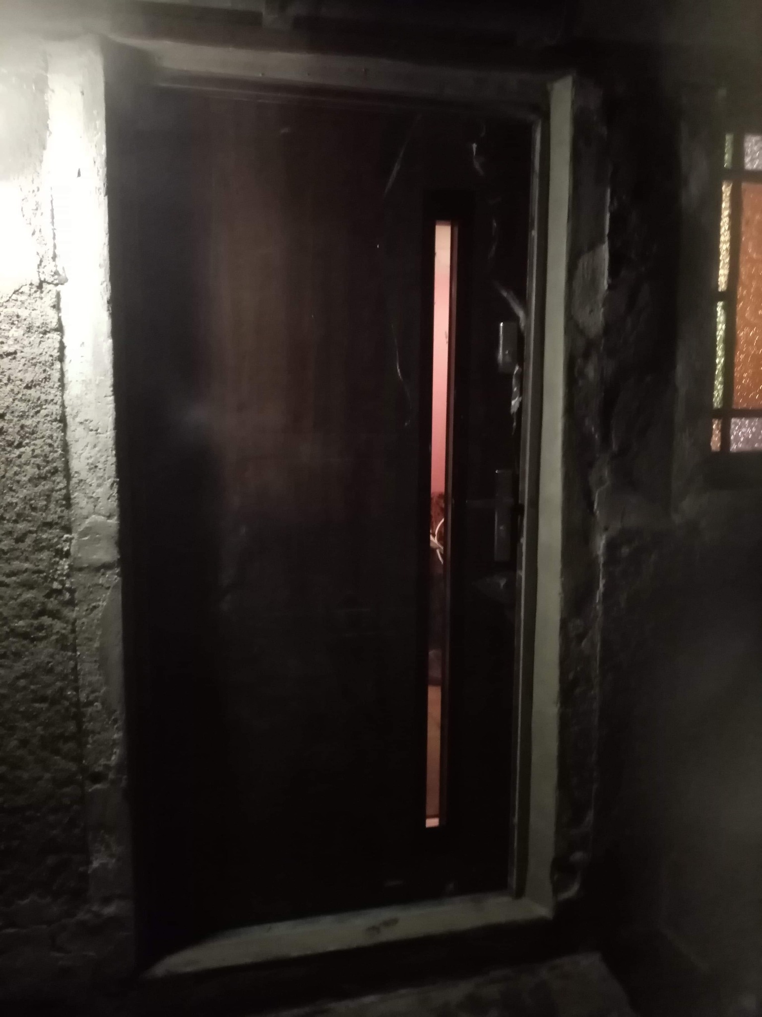 Ciemne, drewniane drzwi wejściowe z wąskim, pionowym przeszkleniem, osadzone w szorstkiej, betonowej ścianie. W tle widoczne okno z kolorowymi szybami.