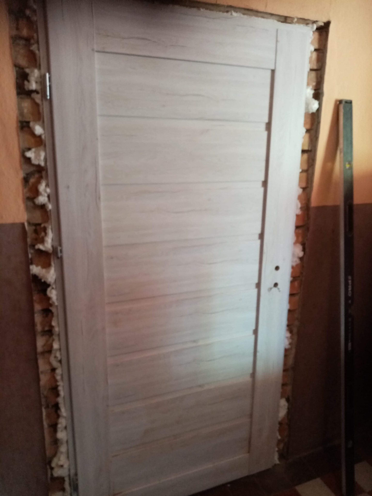 Drewniane drzwi wewnętrzne z poziomymi panelami, osadzone w murze z widoczną pianką montażową i poziomnicą opartą o ścianę.