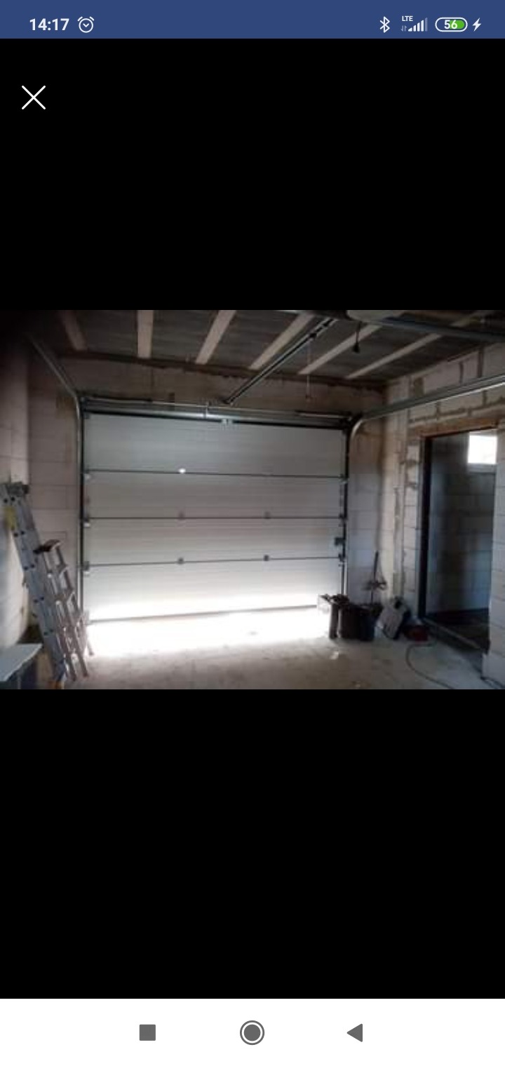 Montaż segmentowej bramy garażowej w surowym wnętrzu, widoczna drabina oparta o ścianę, betonowy strop i otwór drzwiowy obok bramy.