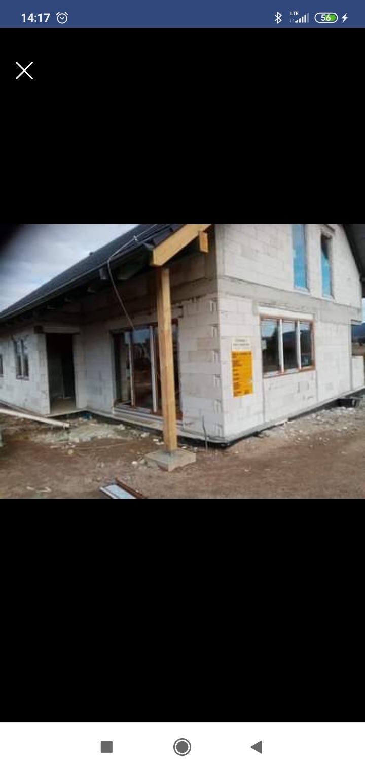 Surowy stan budynku w trakcie budowy z widocznymi oknami i drzwiami, ściany z białego pustaka, drewniane elementy konstrukcyjne zadaszenia, teren wokół budynku niezagospodarowany.