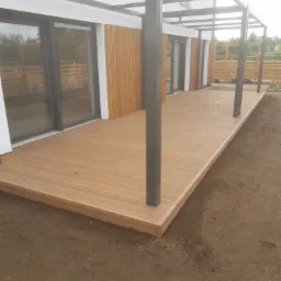 Nowoczesny taras z desek kompozytowych w kolorze jasnego drewna, zadaszony metalową konstrukcją, przylegający do budynku z drewnianą elewacją i dużymi oknami przesuwnymi, otoczony świeżo...