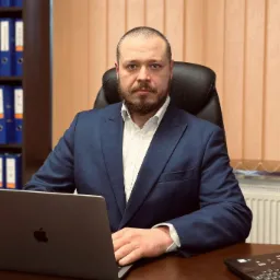 Portret mężczyzny w niebieskim garniturze siedzącego przy biurku z laptopem, w tle regał z segregatorami i beżowe żaluzje.