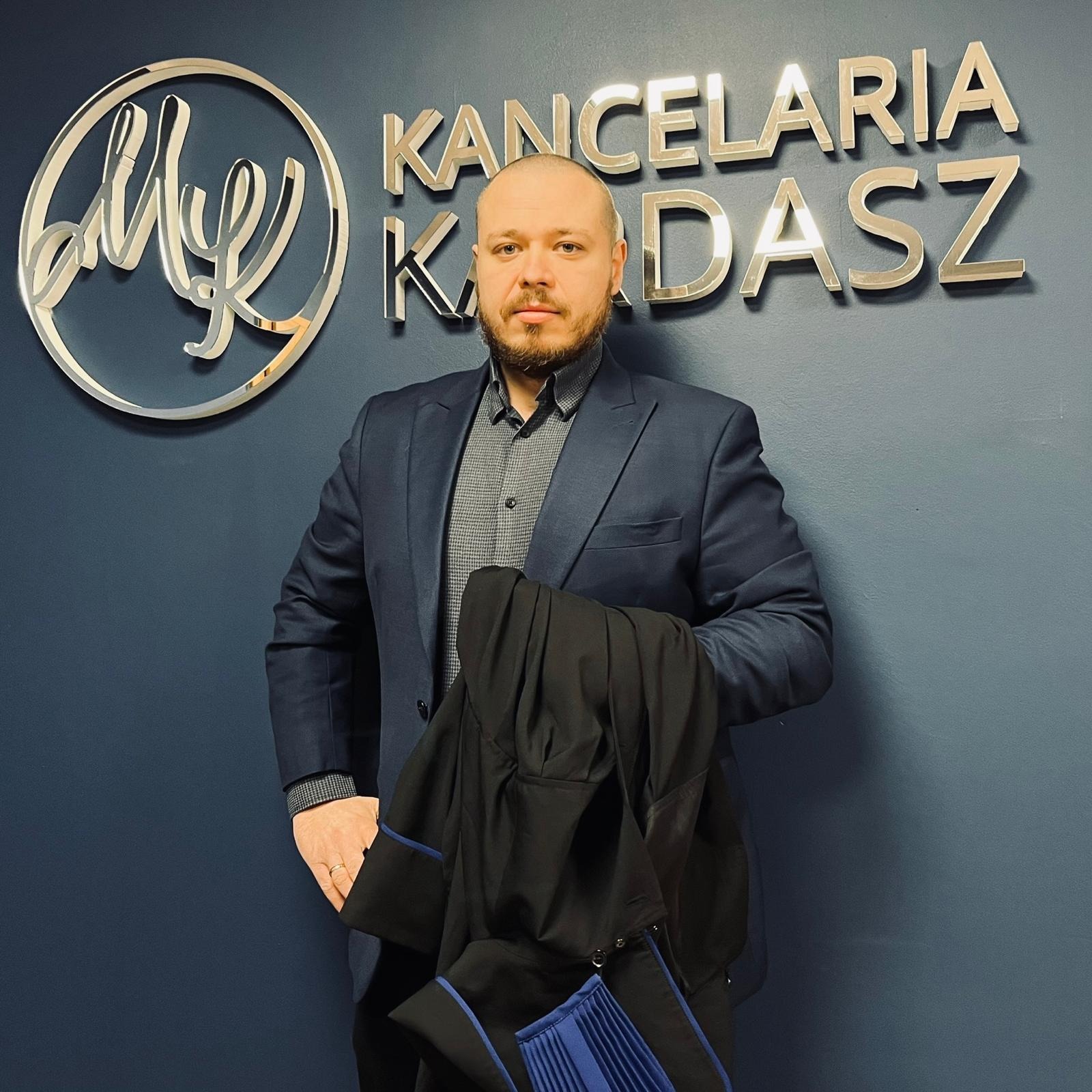 Portret mężczyzny w garniturze trzymającego togę adwokacką na tle ściany z logo kancelarii.