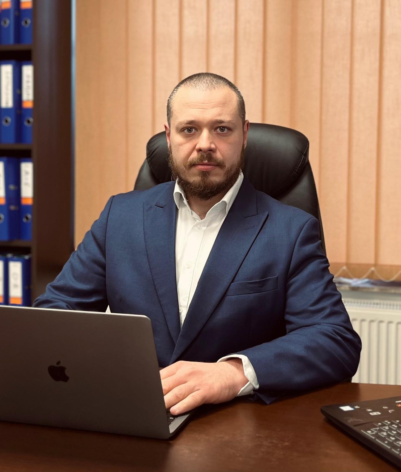 Portret mężczyzny w niebieskim garniturze siedzącego przy biurku z laptopem, w tle regał z segregatorami i beżowe żaluzje.