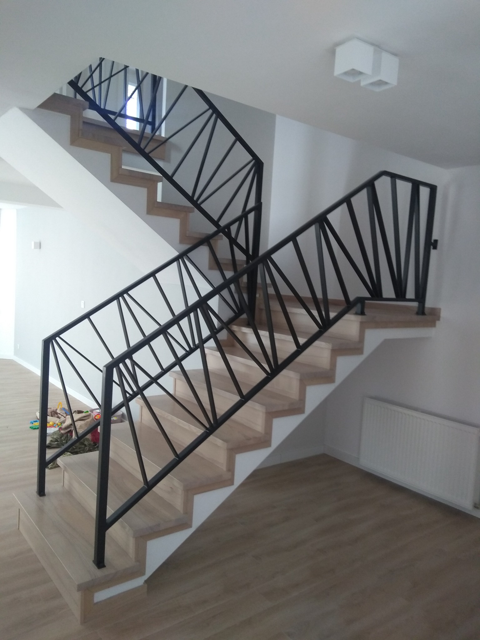 Nowoczesne schody z drewnianymi stopniami i czarną, geometryczną balustradą, widok na dwa biegi schodów w jasnym wnętrzu.