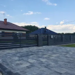 Nowoczesna posesja z przesuwną bramą wjazdową i ogrodzeniem z poziomych, metalowych profili, wspartych na słupkach z szarej kostki betonowej, z widoczną kostką brukową na podjeździe.