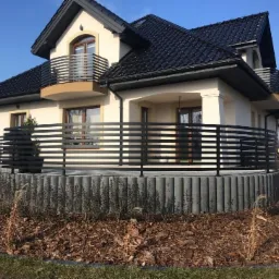 Nowoczesne ogrodzenie poziome z czarnych profili, otaczające taras domu z beżową elewacją i czarnym dachem, posadowione na podmurówce z betonowych bloczków, z widocznym fragmentem ogrodu z korą.