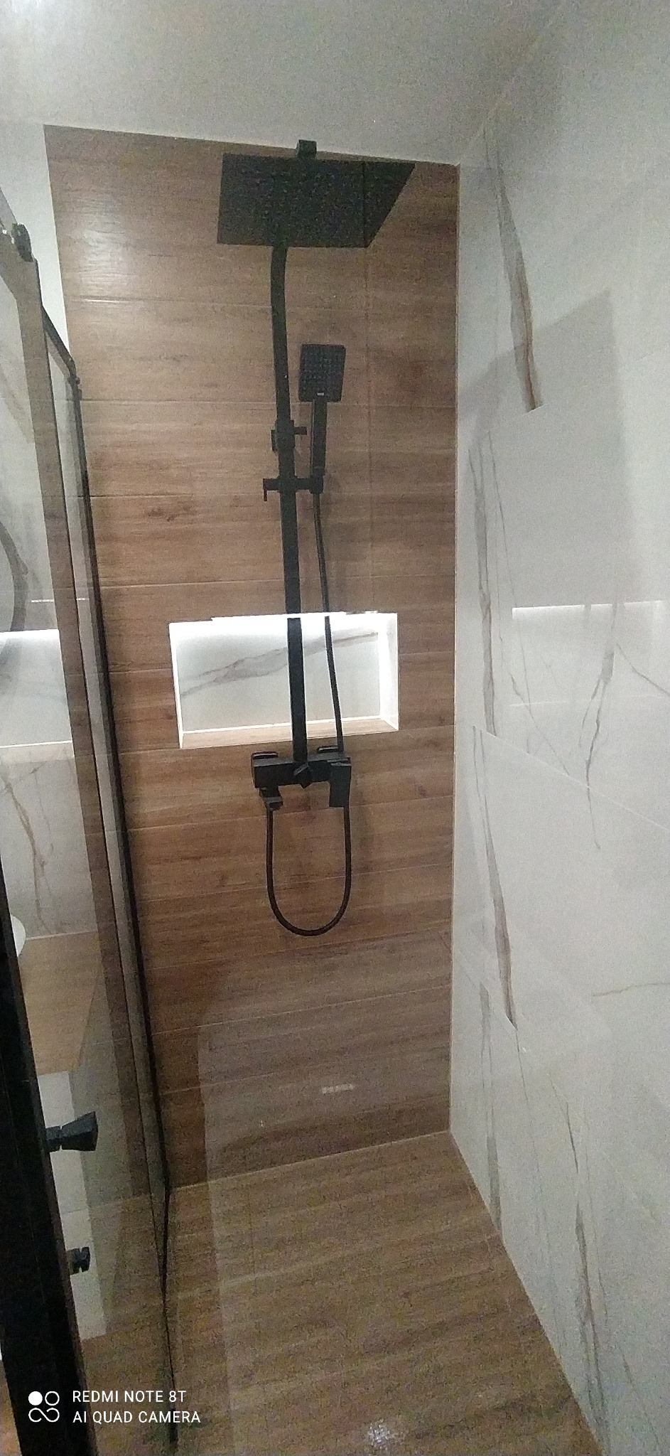 Nowoczesny prysznic z czarną armaturą, drewnopodobnymi płytkami i podświetlaną wnęką na kosmetyki.