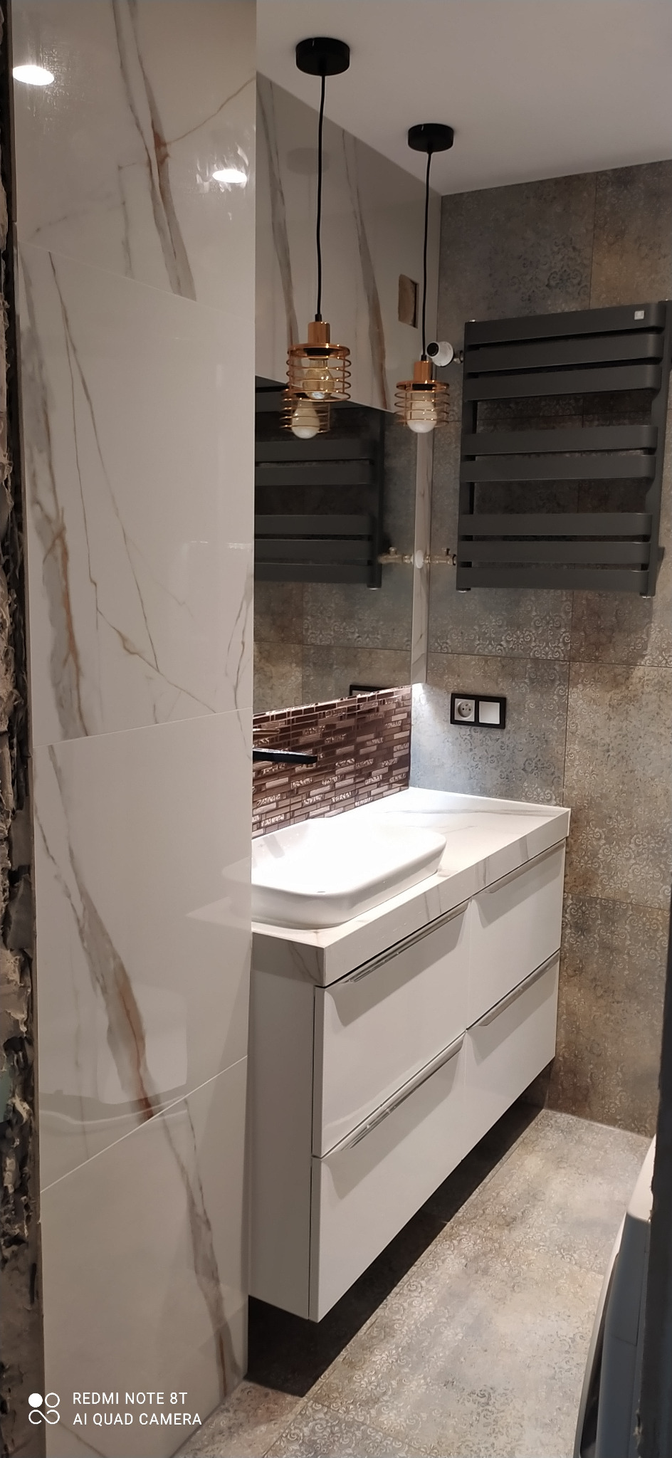 Nowoczesna łazienka z białymi płytkami imitującymi marmur, wiszącą szafką z umywalką nablatową i grzejnikiem drabinkowym w kolorze grafitowym, oświetlona dwoma złotymi lampami wiszącymi.