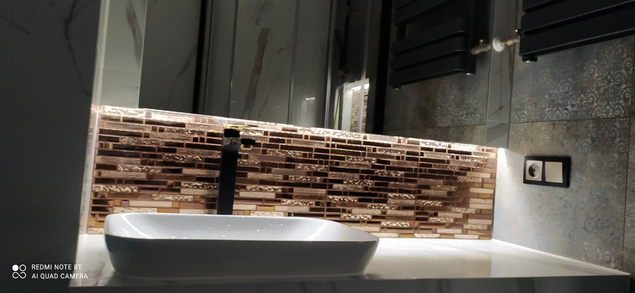 Nowoczesna umywalka nablatowa z czarną baterią na tle ściany wyłożonej mozaiką w odcieniach brązu i beżu, z widocznym gniazdkiem elektrycznym i grzejnikiem drabinkowym w tle.