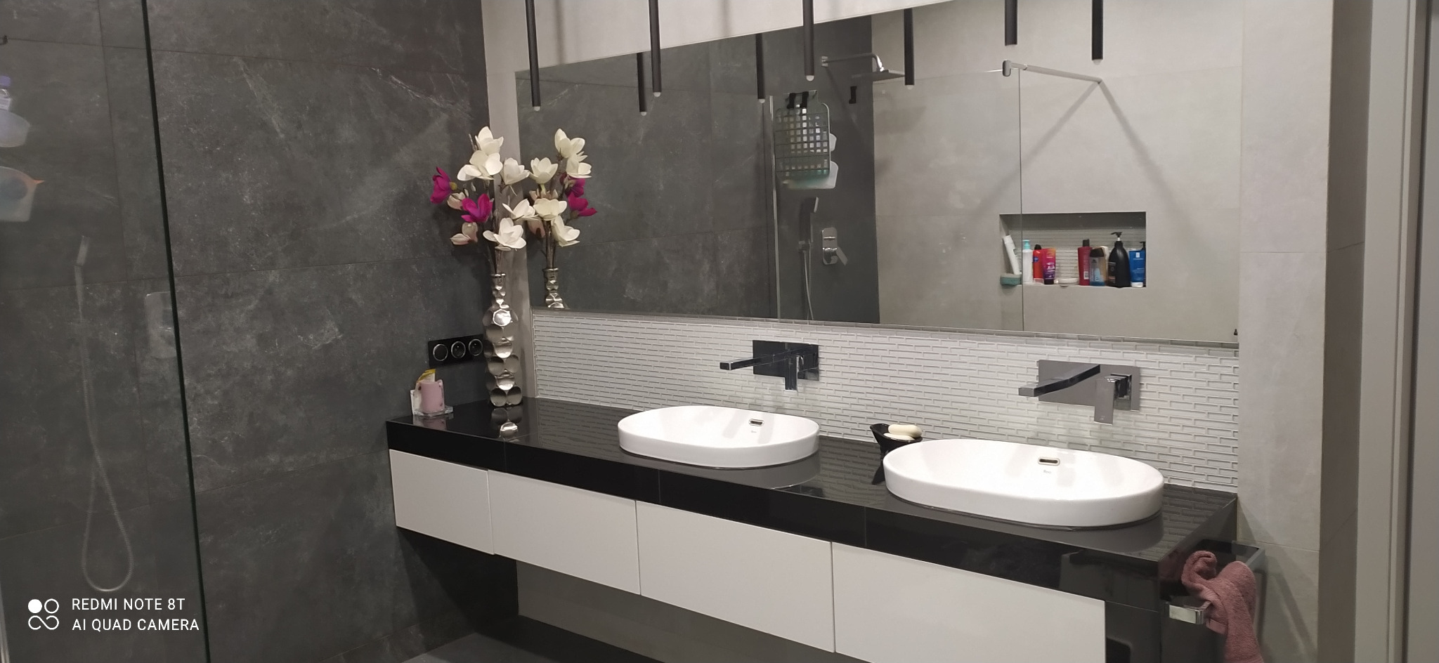 Nowoczesna łazienka z dwoma białymi umywalkami na czarnym blacie, szara ściana z efektem kamienia, duże lustro z widocznym odbiciem kabiny prysznicowej i półką z kosmetykami, srebrny wazon z kwiatami.