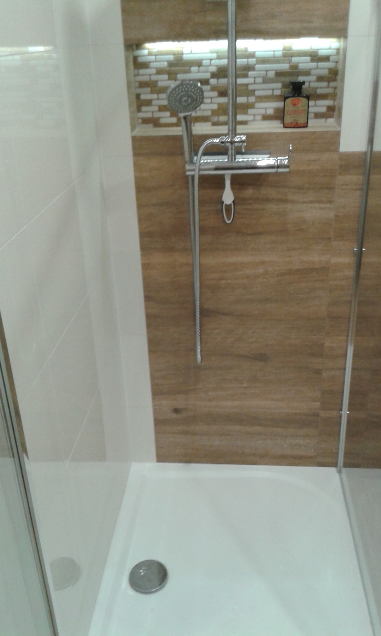 Kabina prysznicowa z brązowymi płytkami imitującymi drewno, mozaika w niszy, bateria prysznicowa z deszczownicą i półką na kosmetyki, butelka kosmetyku Jantar w niszy.