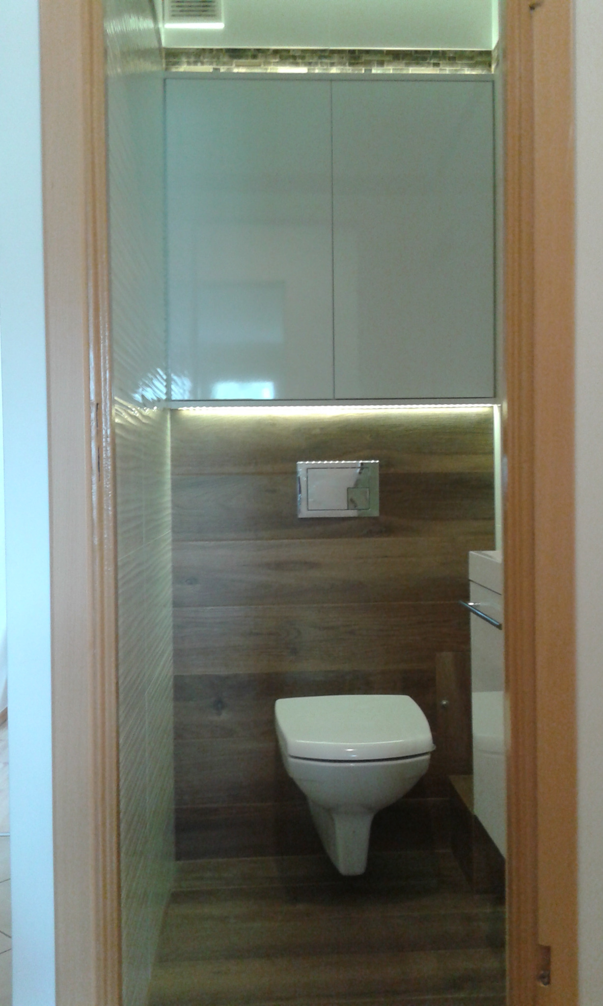 Nowoczesna toaleta z podwieszaną miską WC, drewnopodobnymi płytkami na ścianach i podłodze, oświetleniem LED podszafkowym i szafkami wiszącymi w kolorze jasnym, widziana z perspektywy wejścia.