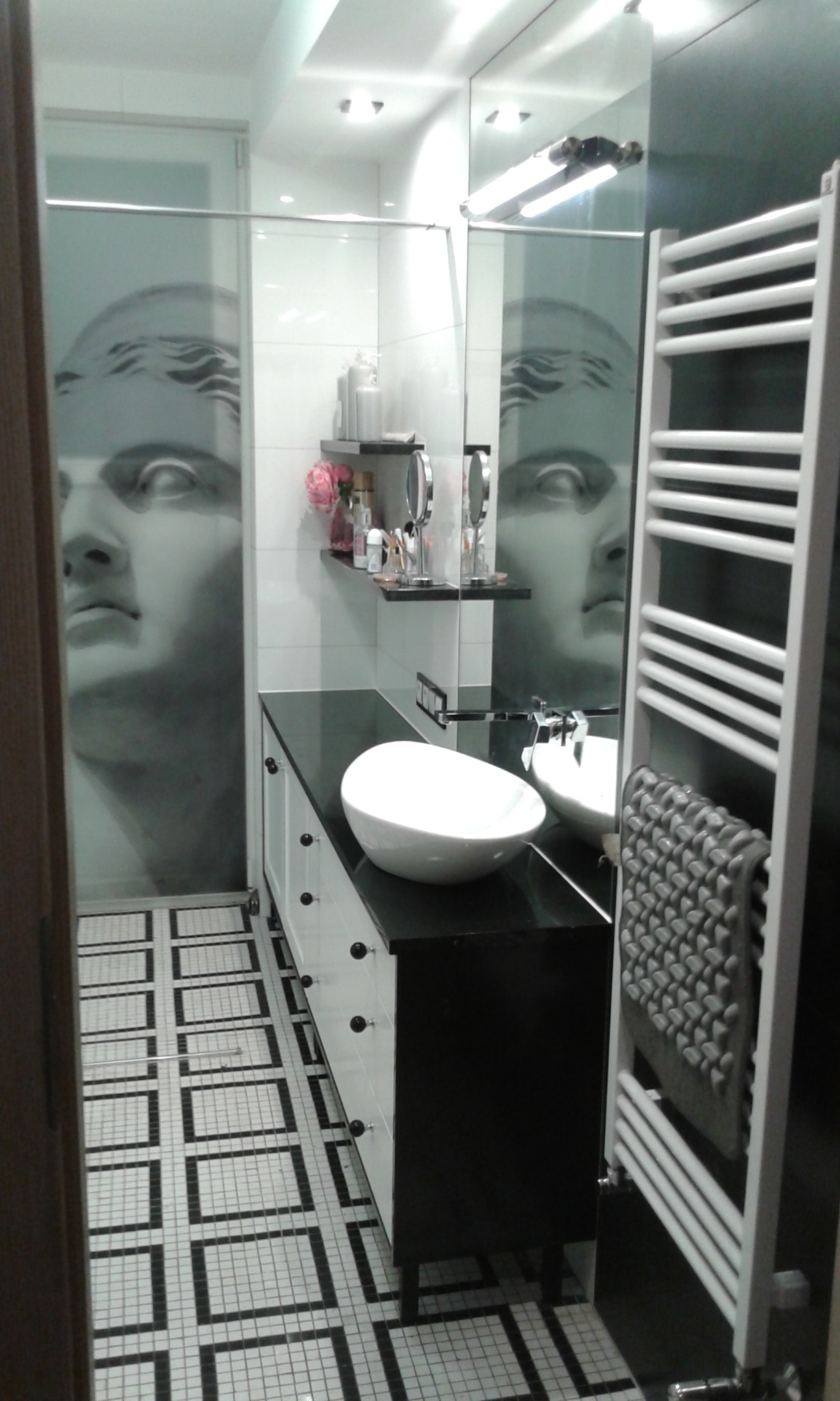 Nowoczesna łazienka z umywalką nablatową na czarnym blacie, białą szafką z okrągłymi uchwytami, grzejnikiem drabinkowym i dekoracyjną grafiką twarzy na szklanej kabinie prysznicowej, dopełniona...