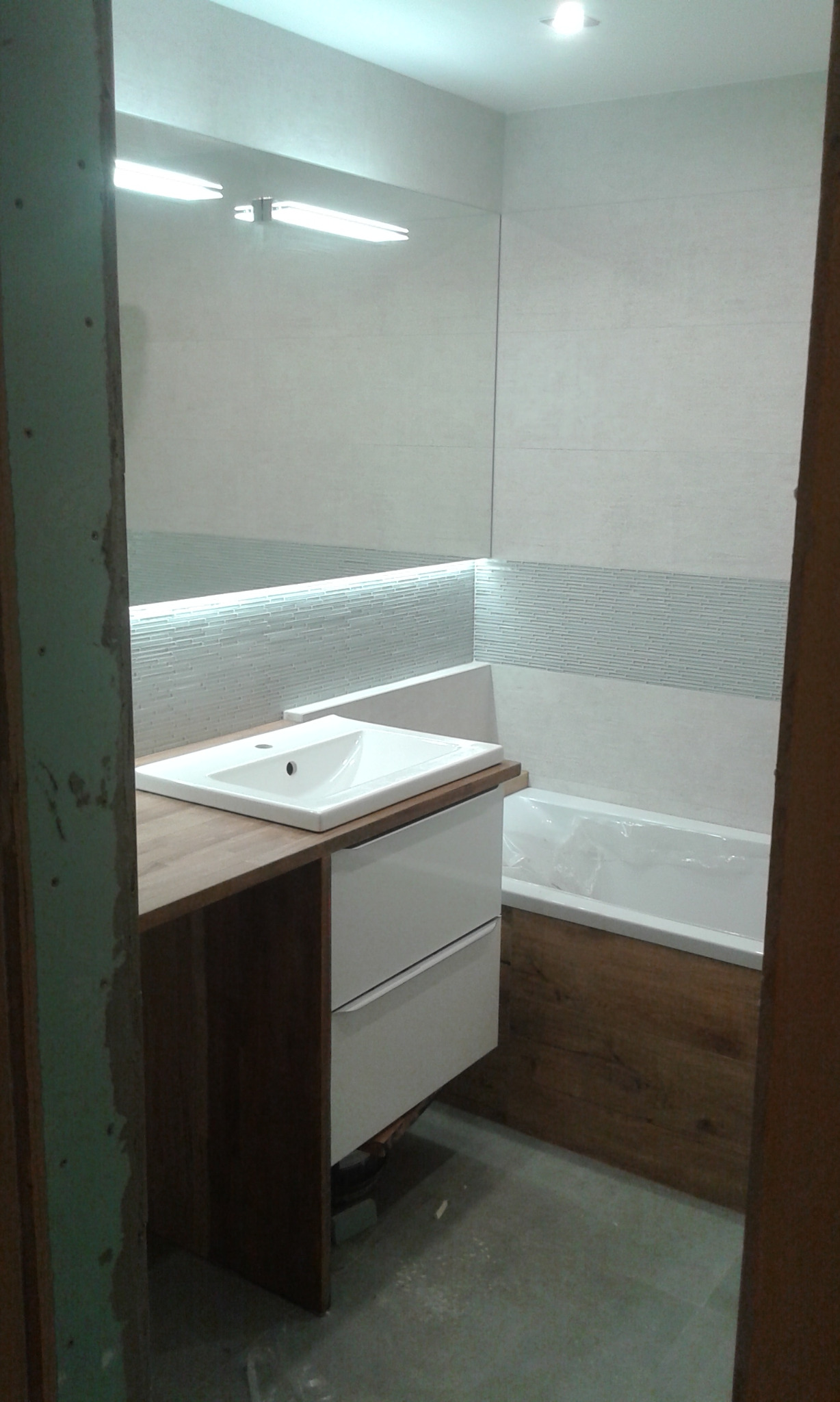 Nowoczesna łazienka z drewnianymi akcentami: umywalka nablatowa na drewnianym blacie, białe szafki z szufladami, wanna obudowana drewnem, lustro z oświetleniem LED, szare płytki na podłodze...