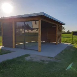 Nowoczesna drewniana konstrukcja z częściowo przeszkloną ścianą, stojąca na wybrukowanym tarasie, otoczona zielonym trawnikiem w słoneczny dzień.